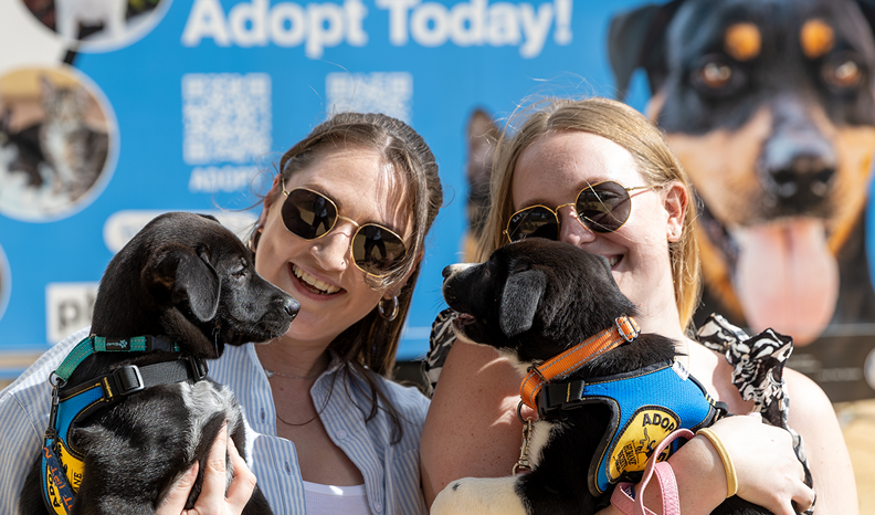 Two young girls each hug an adoptable puppy in front of an Adopt Today sign on a truck